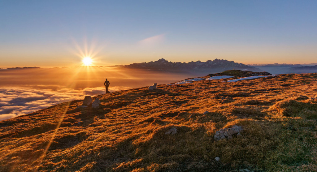 Hiker and a dog at the top of the mountain at sunset with aeautiful view of Julian Alps.