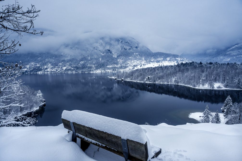 A wooden bench with a view of Lake Bohinj covered in snow the next day after snowfall