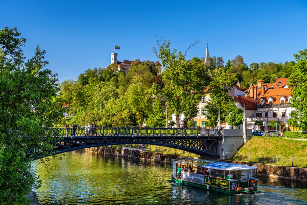 Ljubljanica River with a tourist boat under the bridge and Ljubljana Castle in the background